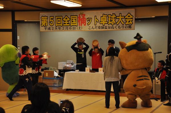 左側にマイクを持った男性が話していて、その中央のテーブルに男女3人が立っていて桶を頭の高さにあげている写真