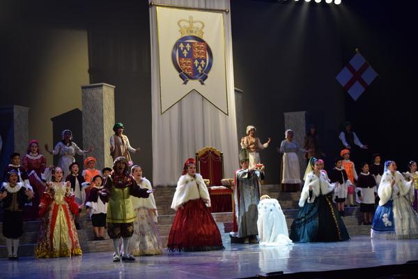 赤いドレスを着た女性の隣に舞台真ん中に王冠をもった男性の前にひざまずいている人がいて周りの出演者の人達が両手で動きを表したり、音楽に合わせて歌っている写真