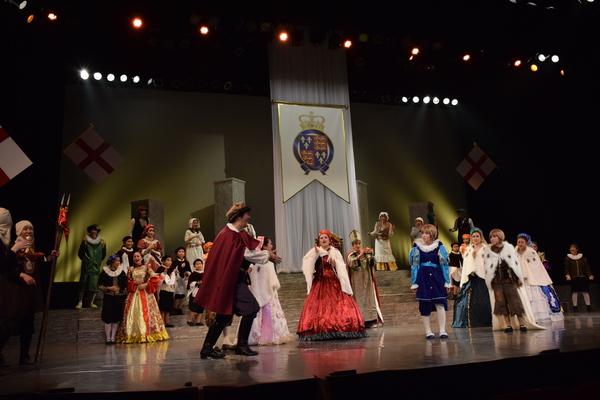 茶色のベレー帽を被り、赤のマントを付けた男性が演技をしながら舞台の真ん中に近づいてくる様子の写真