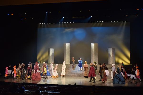 階段の途中にたつ茶色の衣装と青と紫の衣装を着た2人のほうを他の様々な衣装をきた出演者の人達がみている写真