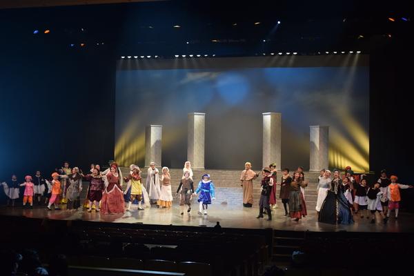 茶色と青と紫の衣装をきた主人公がステージ真ん中に手をつないでたち周りの様々な衣装を着た出演者がステージ上に広がり、音に合わせて歌っている様子の写真