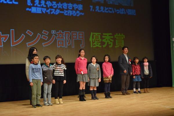 舞台上の後に大きなスクリーンがあり、その前に小学生の児童が8名と赤い花の胸章を付けている男性と女性が立って写っている写真