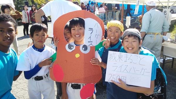 手作りの被り物ライスバーガーから顔を出している男の子と、ライスバーガーの値段が書かれた画用紙を持っている男の子らが一緒に記念撮影している写真