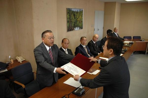 植村 みつる議員がグレーのスーツを着た男性から証書を手渡しでもらっており、横に座っている男性の方々が見守っている様子の写真