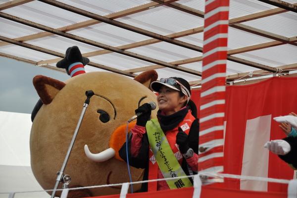 丹波篠山ふるさと大使の熊谷 奈美さんがマイクでお話している隣でマスコット人形が見守っている写真