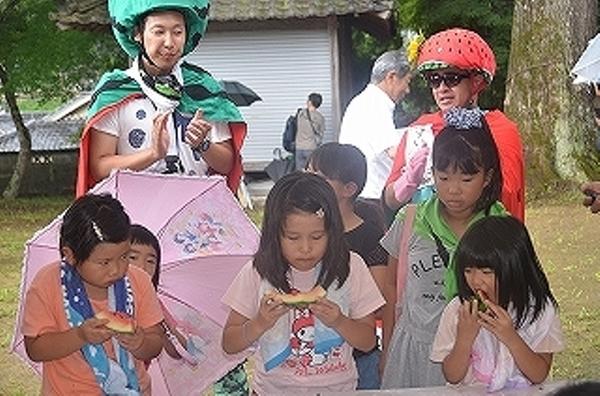 3人の女の子がスイカの早食い競争に参加し3人とも赤い果肉を食べきっており、後ろのスイカの被り物をしている男性2名が手を叩いて応援している様子の写真