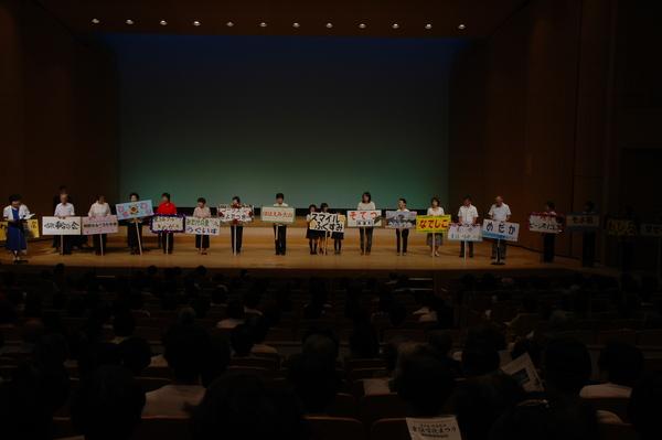 舞台の上に「うぐいす」、「そてつ」などの文字の書かれたプラカードを持った人たちが横一列に並んでいる写真