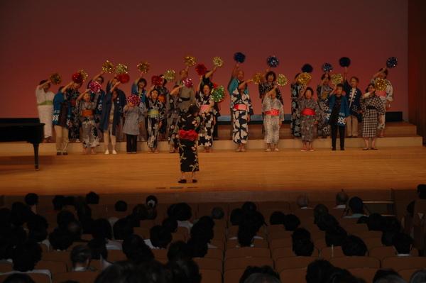 舞台の上で浴衣を着た人達がキラキラしたボンボンを持って上に大きく手を振っている写真