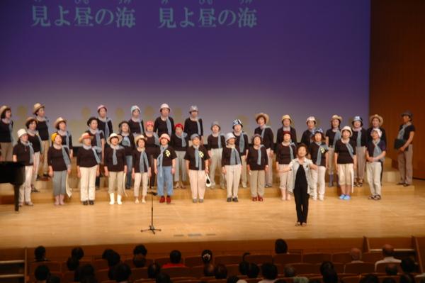 ベレー帽をかぶって首にスカーフを巻いた女性たちが舞台の上で歌っている写真