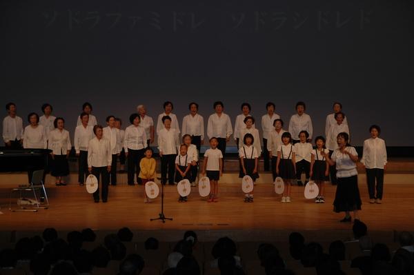 20名ほどの白いシャツを着た女性たちの前に7名の小学生が立っていて全員で歌を歌っている写真