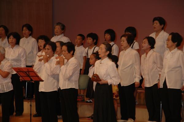 女性や小学生たちが歌っていて、その中央で二人の女性がオカリナを吹いている写真