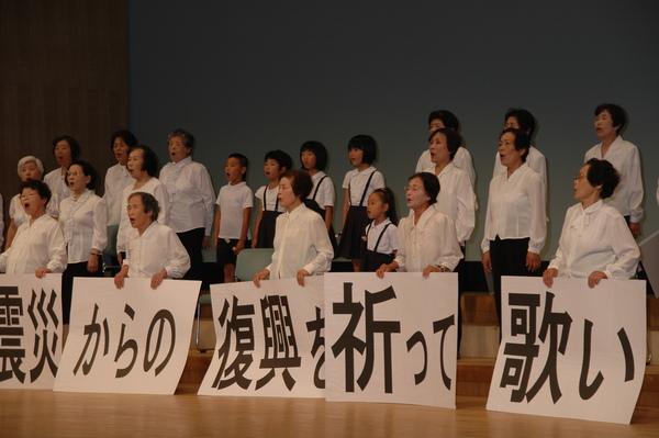 「震災からの復興を祈って歌い」と書かれた紙を持った女性たちが歌っている写真