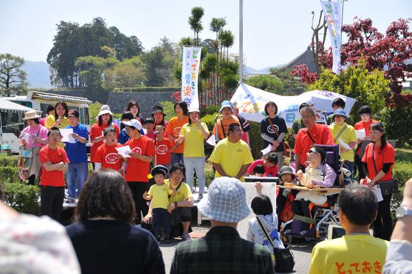 オレンジ、黄色、青色のTシャツを着た方々が前に立ち、歌を歌っている、その様子を見ている周りの方の写真