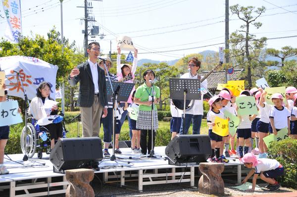 ステージ上で歌を歌っている男女とその横にピンクの帽子を被った保育園児が文字の書かれた紙を持っている写真