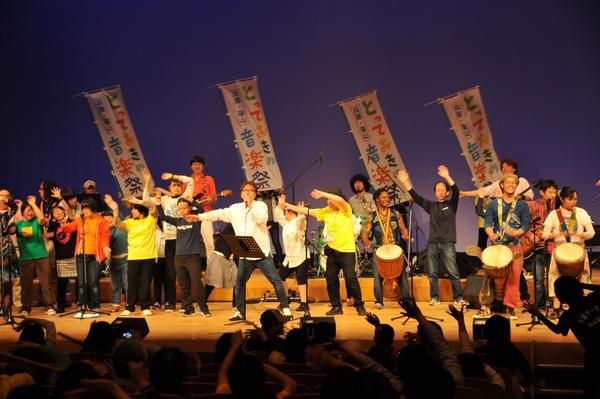 マイクを持ち、歌を歌っている男性の歌に合わせて両手を広げ踊っている人や太鼓を叩いている方々の写真
