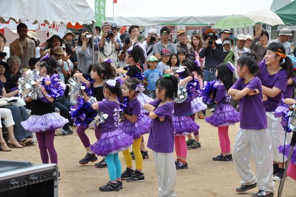 紫色の衣装とボンボンを身に着けているダンスチームの子ども達が、両手を胸の前に組んでポースを決めダンスを踊っている様子の写真