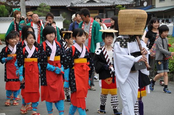 虚無僧が天蓋を被り尺八を吹いており、その後ろに茶摘み娘の衣装を着た女の子達が後をついて歩いている様子の写真