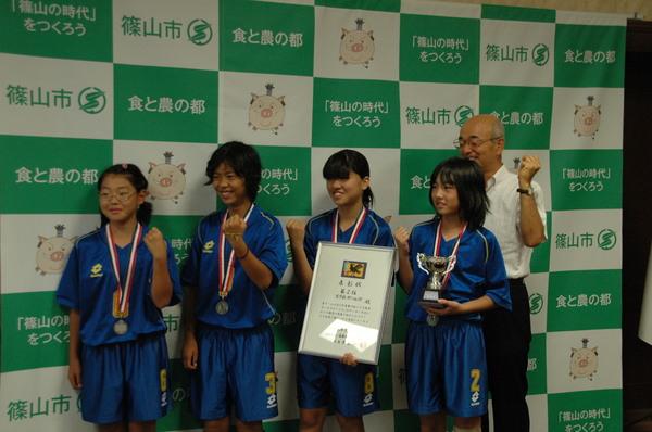 青いユニホームを着た女子4名のうち一人が2位の表彰状を持ち、一人がトロフィーを持ってガッツポーズをしており、その後ろに市長が立ってガッツポーズをしている写真