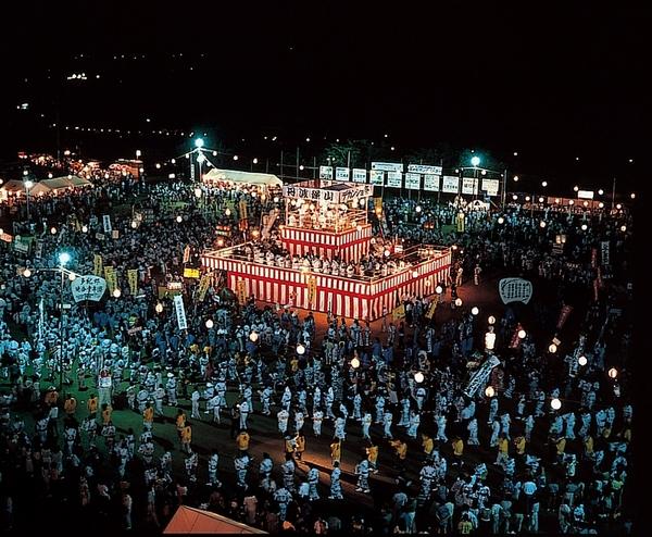 夜のお祭りで紅白幕の舞台に提灯が照らされ沢山の人が踊っている写真