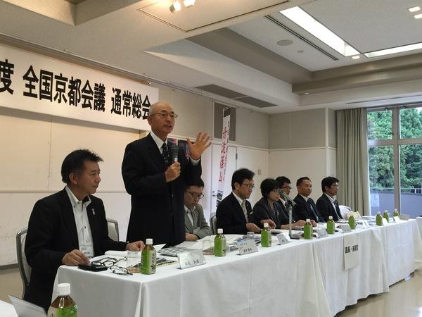 8名のスーツを着た男女がテーブルに座っていて、市長が立ってマイクで話している写真