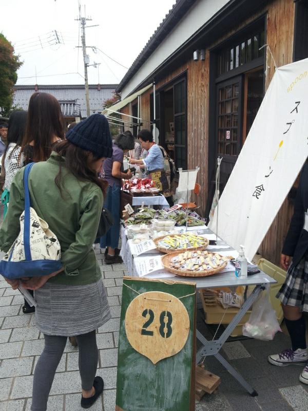 28と書かれた木の看板が置かれた店前の机に並ぶ、食品を興味深く見ている女性の写真