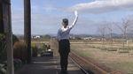 駅のホームで白いシャツを着た女性の名物ボランティア駅長さんが右手を上げて立っている後ろ姿のシーンの写真