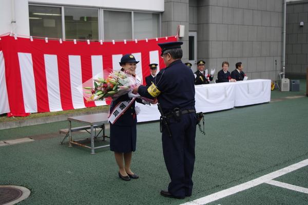 駐在所のご主人から一日署長を務める奥様の瀬頭 とよみさんへ花束が贈呈されているのを来賓者が見ている写真