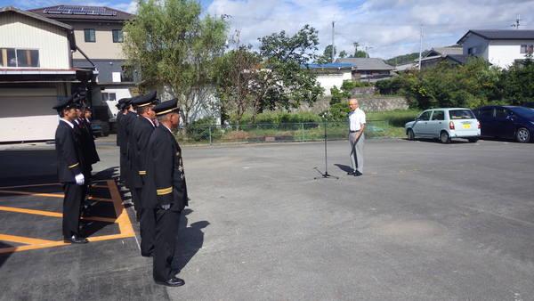 消防詰所の横で市長が挨拶をしているのを制服姿の消防団員らが聞いている写真