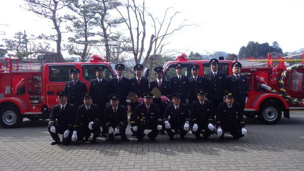消防車両2台の前で、前列と後列に整列する制服姿の男性消防団の写真