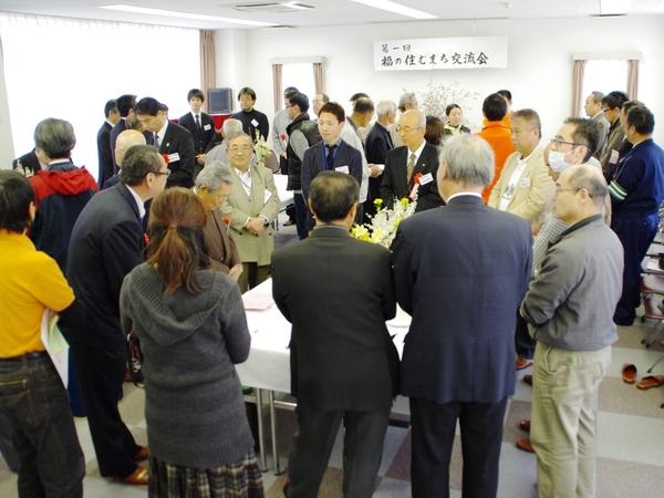 第一回 福の住むまち交流会の横断幕が掛けてある部屋にテーブルがいくつか設置されており、各テーブルを出席者が囲んで話をしている様子の写真