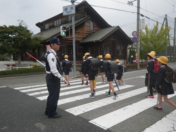 ランドセルを背負い、黄色い帽子をかぶった小学生たちを、横断歩道で警察官が誘導している写真