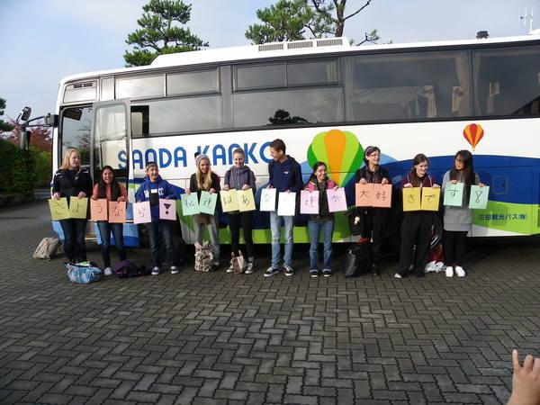 留学生10人がバスの前で文字の書いた紙を一人一人両手に持ち立っている写真