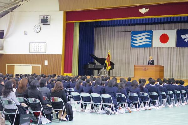 生徒、保護者がステージに立っている男性の話を真剣に聞いている様子を右斜め後ろから写した写真