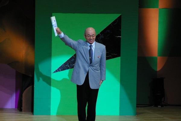 市長が舞台の上で、右手に冊子を持ち右手を上げて写っている写真