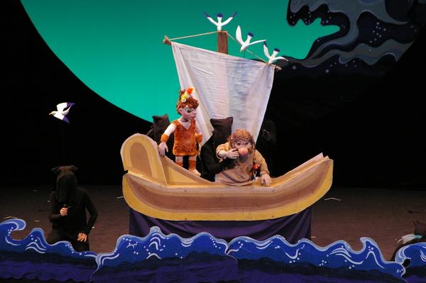 海の上に小さな帆船が浮かんでおり、船には主人公の少年ナギと茶色い衣装を着た人形が乗っている火の鳥の公演の一場面の写真