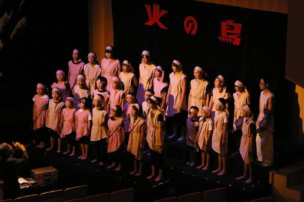 「火の鳥」と赤い文字で書かれた幕のまえで、頭に白いハチマキを巻き、白い衣装を身に着けた子ども達が、3列に並んで歌を歌っている写真