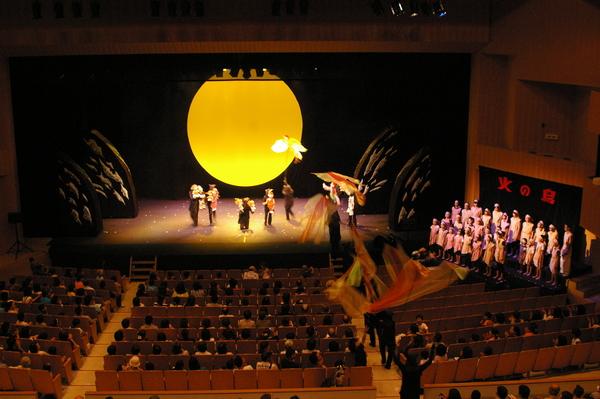 舞台の上には大きな黄色い満月が写しだされ、会場内を火の鳥がとんでおり、登場人物の人形と黒子が舞台に立っており、右側には白い衣装を身に着けた合唱団が立っており、客席には沢山のお客さんが座っている様子を後方から写している写真