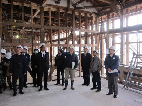 木組みの建物の中で薄い黄色のヘルッメトを被った男性が、関係者の前で、指をさして話をしている写真