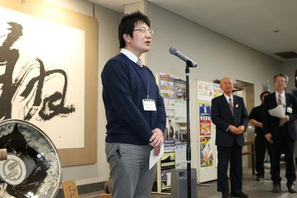 スタンドマイクの前に立ち挨拶をしている山本さんと横で見守る酒井市長と職員の写真