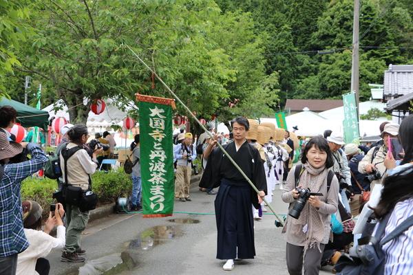 「大国寺と丹波茶まつり」の旗を竹につるして行列の先頭を歩いている袴を着た男性と、沿道の両脇に沢山の観光客がカメラを構えている様子を写している写真