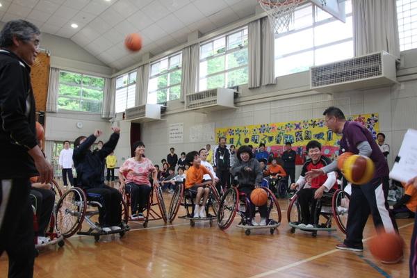車いすに座っている子供たちが、ボールを手に持つ男性を囲み、楽しそうにバスケットボールを投げている写真