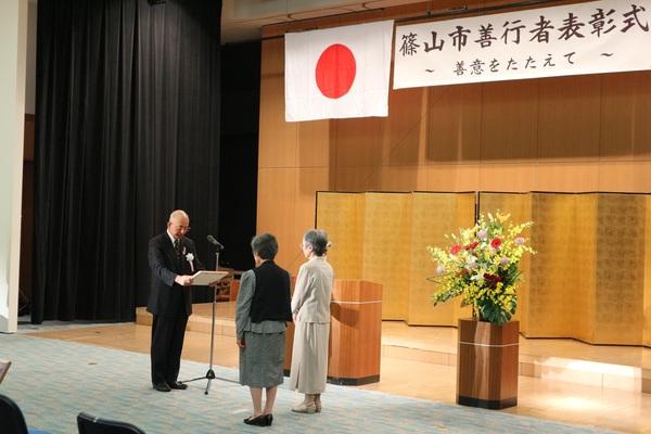 金屏風と綺麗な講壇花の前で、市長より2人の女性が表彰状を読み上げてもらっている様子の写真