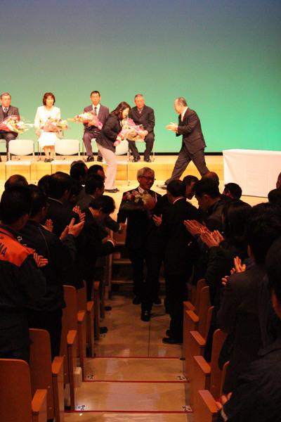花束を受け取った退職者が壇上から下り沢山の人の拍手で送られている写真