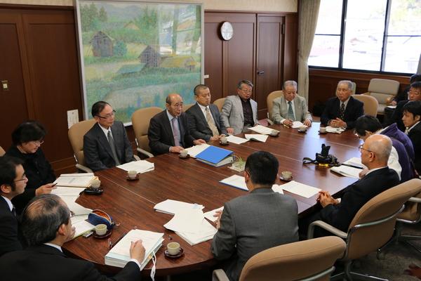 テーブルに書類を置き、スーツを着た男性が10人ほど熱心に議論している写真