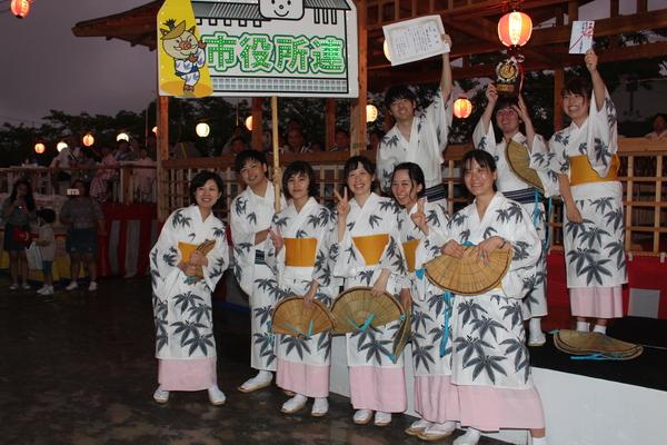 篠山市役所連のプラカード、トロフィ、賞状、賞金を持った男女が、えがおで並ぶ集合写真