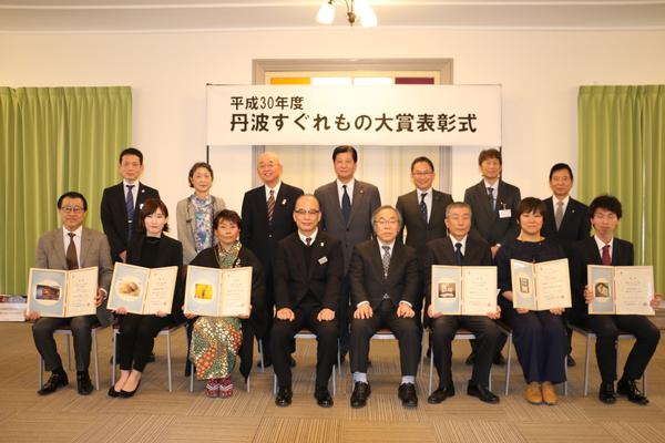 「平成30年 丹波すぐれもの大賞表彰式」の横断幕の前で、受賞者が額を持って座っており、市長及び関係者が一緒に写っている集合写真