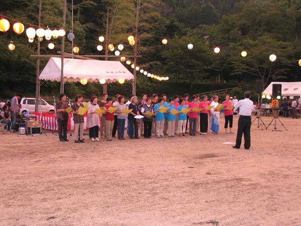 舞台の下で女性達が黄色い紙を持ち指揮者の方を向いて歌っている写真