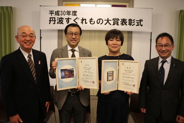 「平成30年 丹波すぐれもの大賞表彰式」の横断幕の前で、市長と表彰の額を持っているアートフェスティバルの中西さん、ビーファームの松村さんと小西県議会副議長の4人が写っている写真