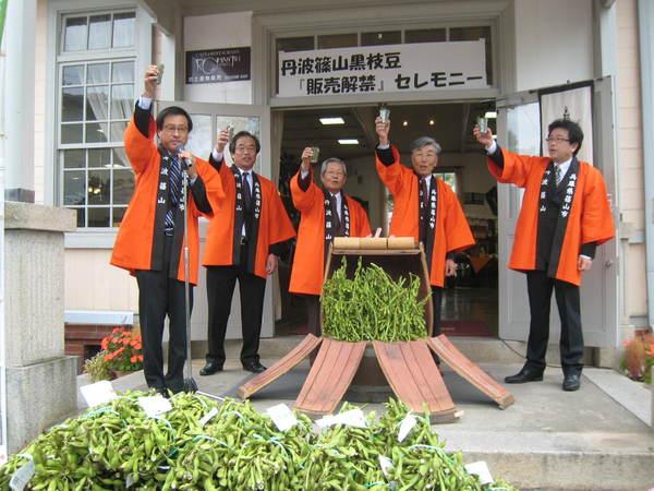 樽や樽の前にたくさんの黒枝豆があり、その後ろにオレンジの法被の男性が右手にコップを持って乾杯している写真