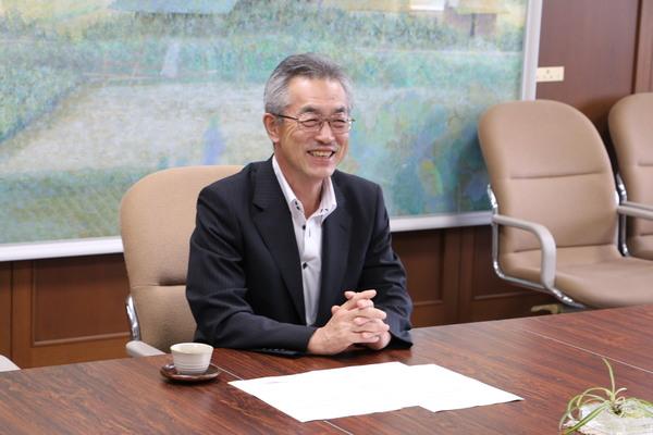 検討委員会の小野 雅之委員長が机の上で両手を組み笑顔で写っている写真